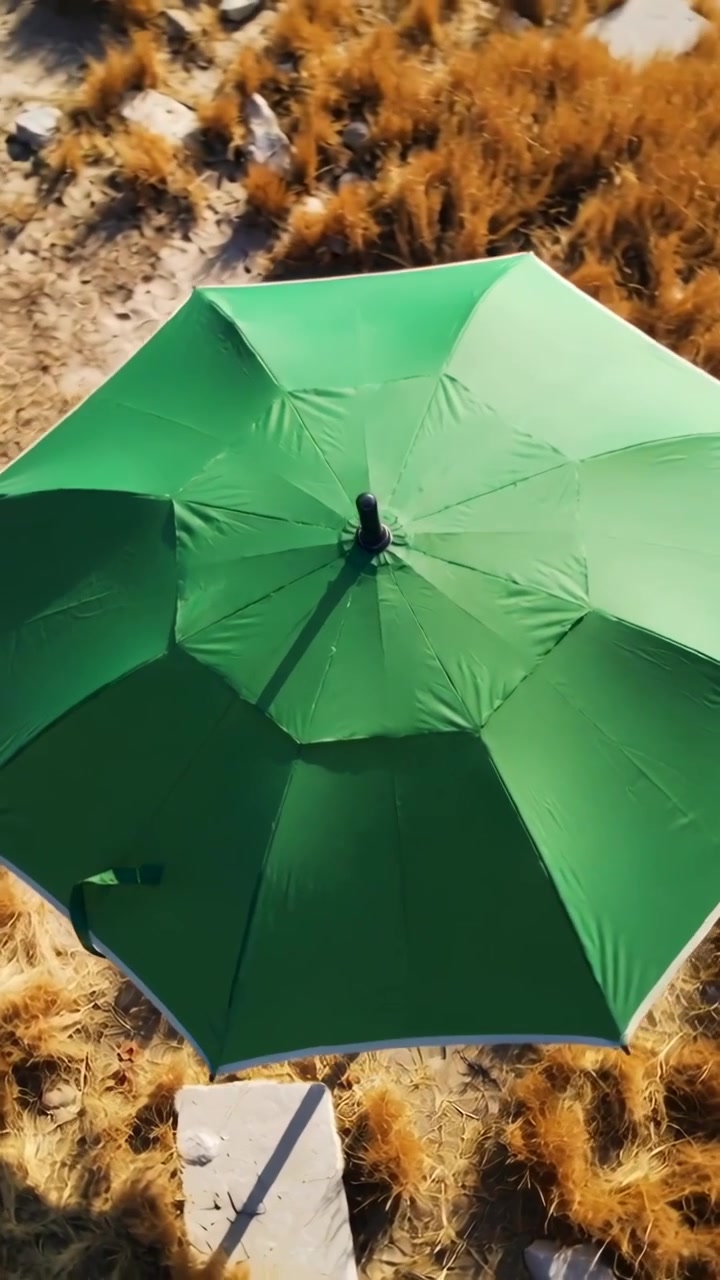 Vertikal video: Drone trækker sig tilbage og afslører en sandklit for at vise en centreret grøn parasol, stang og sten.