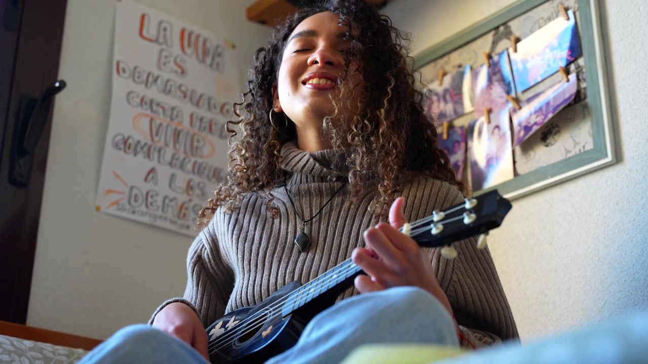 Jovem mulher feliz com cabelo encaracolado tocando ukulele e cantando uma música em seu quarto aconchegante em casa.