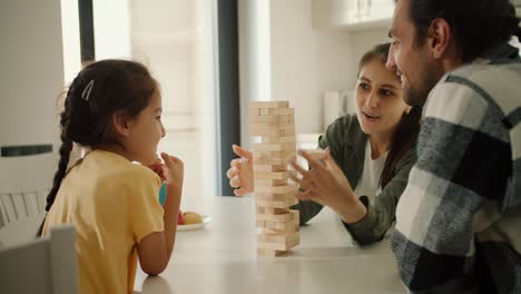 Happy-young-family-brunette-girl-in-a-green-jacket,-her-husband,-brunette-guy-in-a-checkered-shirt-and-their-little-daughter,-brunette-girl-in-a-yellow-t-shirt,-play-Jenga,-a-game-for-developing-finger-motor-skills,-in-a-modern-kitchen-in-an-apartment