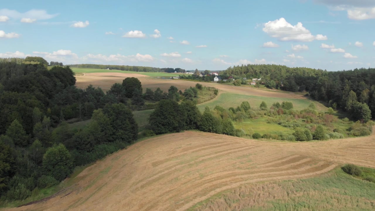 Aerial drone view over rolling farm fields, forest edges and small rural settlements in Kashubia, Poland on a bright summer day with blue sky and clouds.
