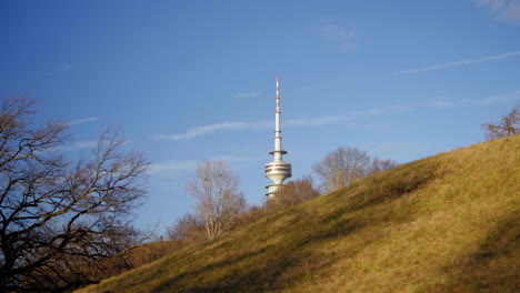 the olympic tower rises above the hilly landscape of olympiapark in munich, germany. captured on a sunny spring day, the scene features green slopes, bare trees, and a clear blue sky.