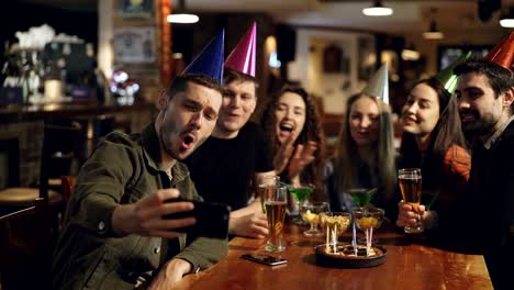 jóvenes felices con sombreros de fiesta de cumpleaños están grabando video con teléfonos inteligentes sentados en la mesa en el bar. están posando, riendo y aplaudiendo.