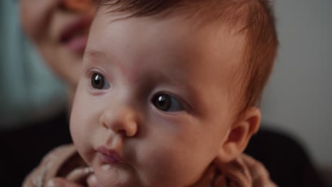 close up of a little baby girl with big eyes looking at the camera and posing in a modern apartment sitting in her mother arms