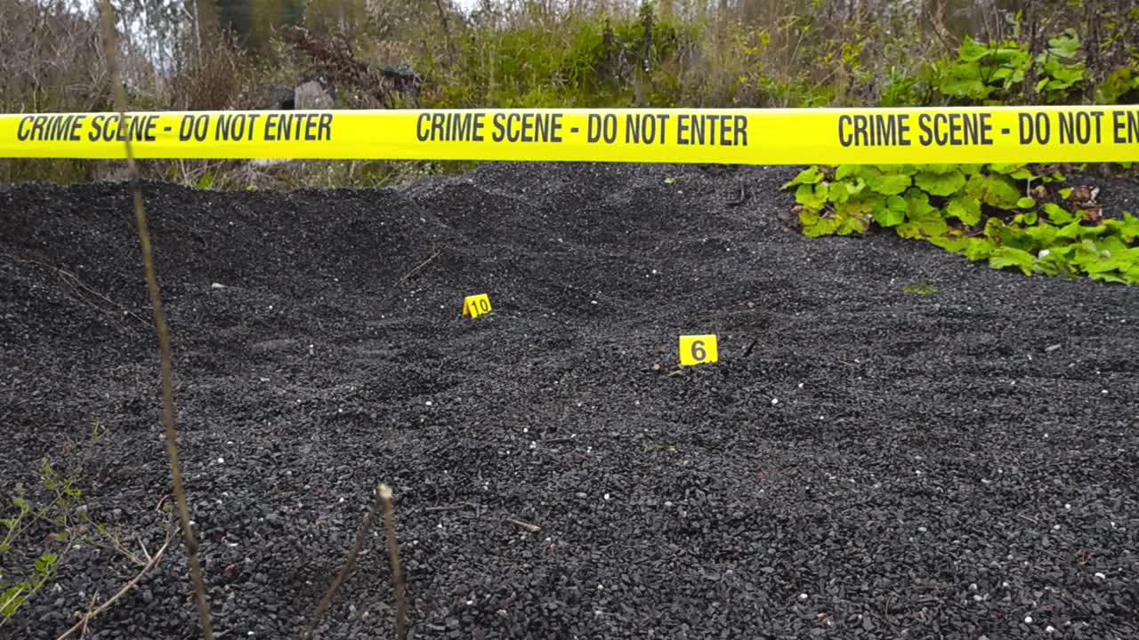 Forensic yellow colored police line or crime scene investigation tape pulled infront of a nature abandoned area with gravel where crime evidence numvered markers are next to evidence. Trees and bushes