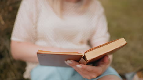 close-up of woman flipping pages in open book while reading outdoors, soft focus on blue shorts and delicate fingernails, peaceful moment of study and reflection with natural blurred background