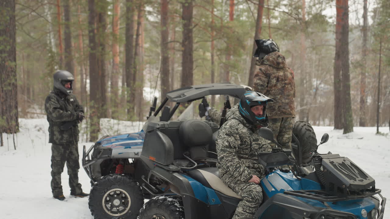 Piloti di ATV nella foresta innevata si radunano attorno a un UTV blu, l'equipaggio con casco prepara il percorso, controllando l'attrezzatura e l'acceleratore, giacche mimetiche e occhiali, pini e neve che cade creano un'atmosfera di spedizione vivace e tesa.