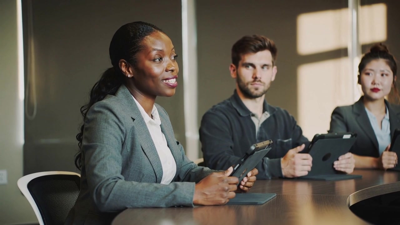 Speaking African American woman in grey blazer holding tablet, presenting to team at curved table