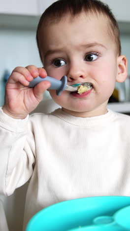 small baby boy eating food from spoon himself. charming kid has some porridge for breakfast. close up. vertical video