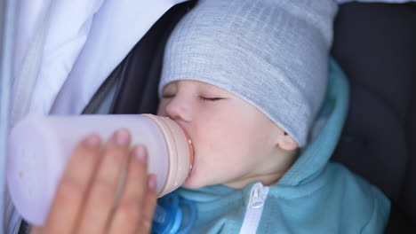 beautiful child wearing grey cap and blazer drinking milk from bottle. healthy baby boy closed his eyes to fall asleep. close up.