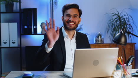 businessman working on laptop smiling friendly at camera and waving hands gesturing hello at office