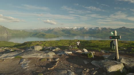 Young-couple-hiking-in-Norway's-Senja-Husfjellet-with-hiking-poles,-snow-capped-peaks-in-background