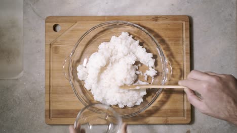 top down view on kitchen counter with a glass bowl of sushi rice and vinegar being spread with a wooden spoon in the dish as part of a recipe