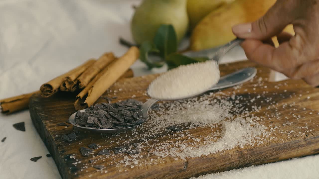 Su un tavolo con una tovaglia bianca ci sono pere, bastoncini di cannella e un tagliere di legno con due cucchiai pieni di zucchero e gocce di cioccolato
