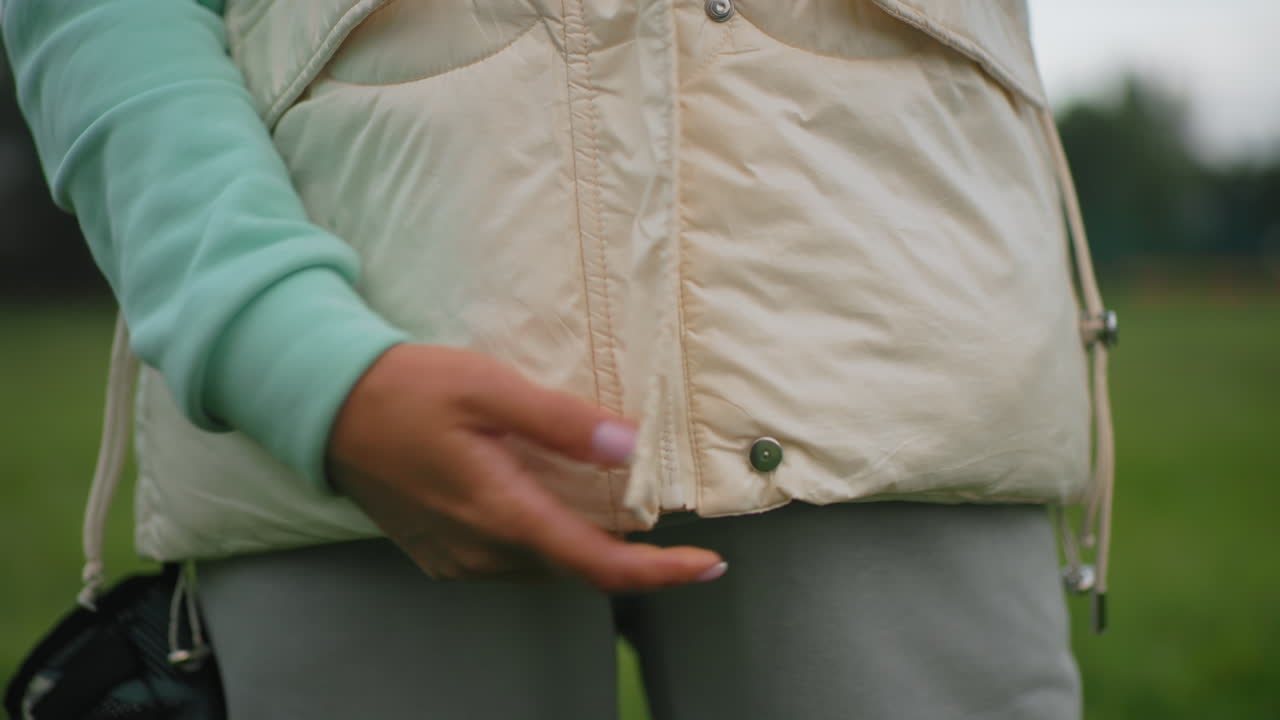 Close up of female trainer zipping up jacket outdoors in grassy field, wearing mint green hoodie and gray sweatpants, preparing for outdoor exercise on cool cloudy day with calm and focused energy