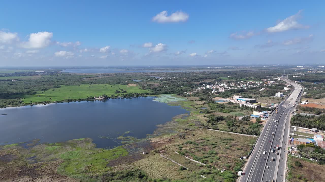 Aerial video of a major highway construction project near Chennai, with new sections and traffic on existing roads. Residential areas, fields, and a large lake are visible, highlighting urban growth