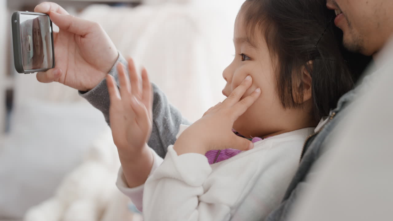 아시아의 아버지와 자녀가 스마트폰을 사용하여 비디오 채팅을 하고 있습니다. 작은 소녀는 주말을 공유하고 있습니다. 아빠와 함께 휴대폰으로 친구 4k와 채팅을 즐기고 있습니다.