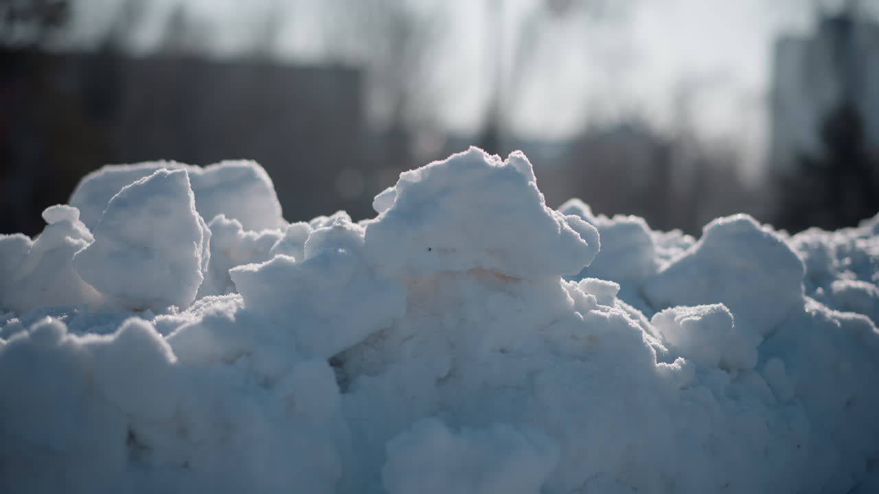 sunlit snow drifts glistening on urban landscape with blurred background of winter trees and buildings under soft daylight closeup of frosty texture and sparkling crystalline mounds