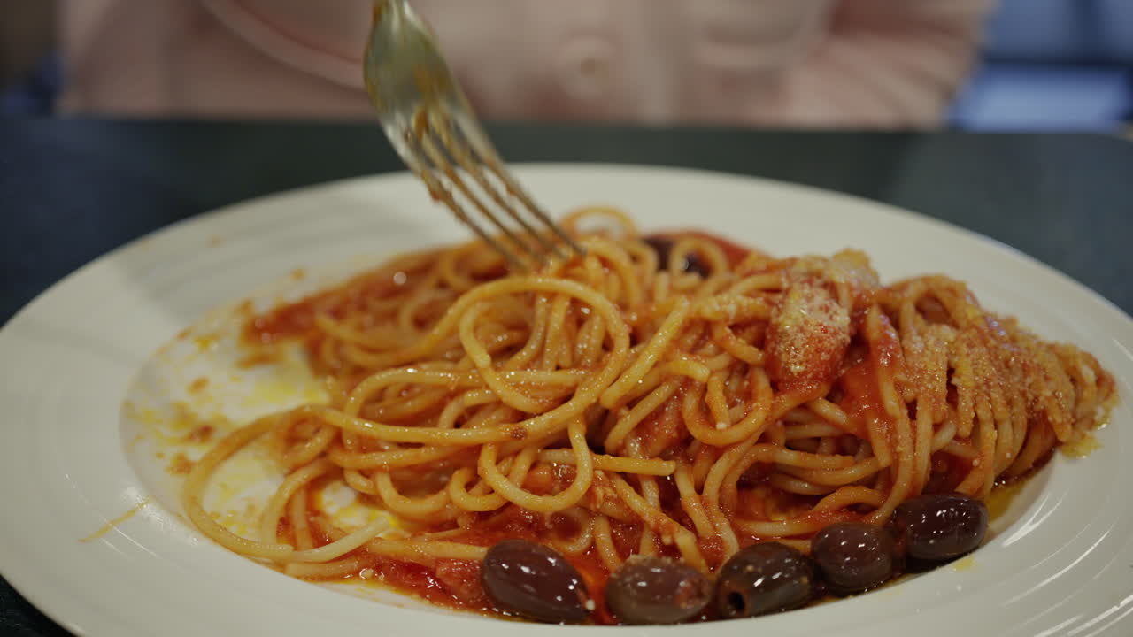 Primo piano di una persona che avvolge spaghetti alla puttanesca con una forchetta, mostrando un ricco sugo di pomodoro, olive e formaggio grattugiato