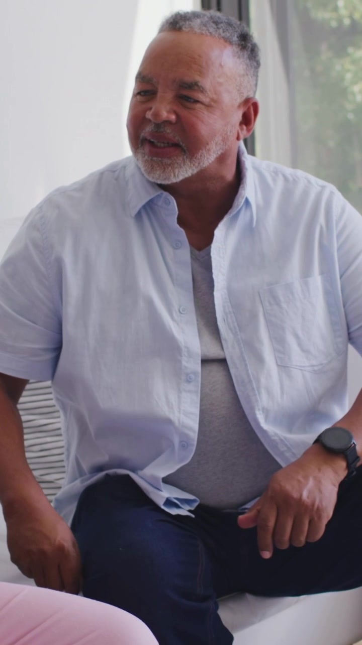 Vertical video: Senior man leaning after cue, smiling on white sofa, wearing blue shirt and watch