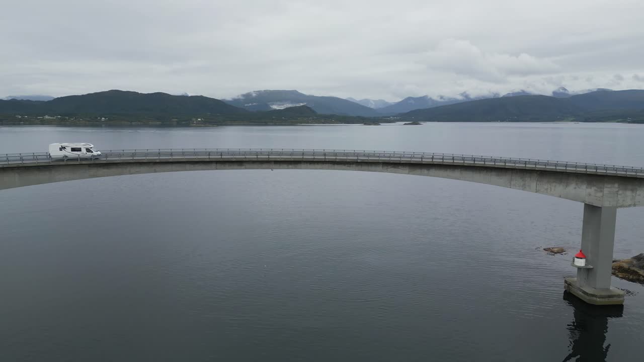좁은 길을 지나는 자동차를 드러내고 다리 끝에 섬을 보여주는 넓은 바다 위로 노르웨이의 가까운 대서양 도로 교량에서 멀리 날아갑니다.