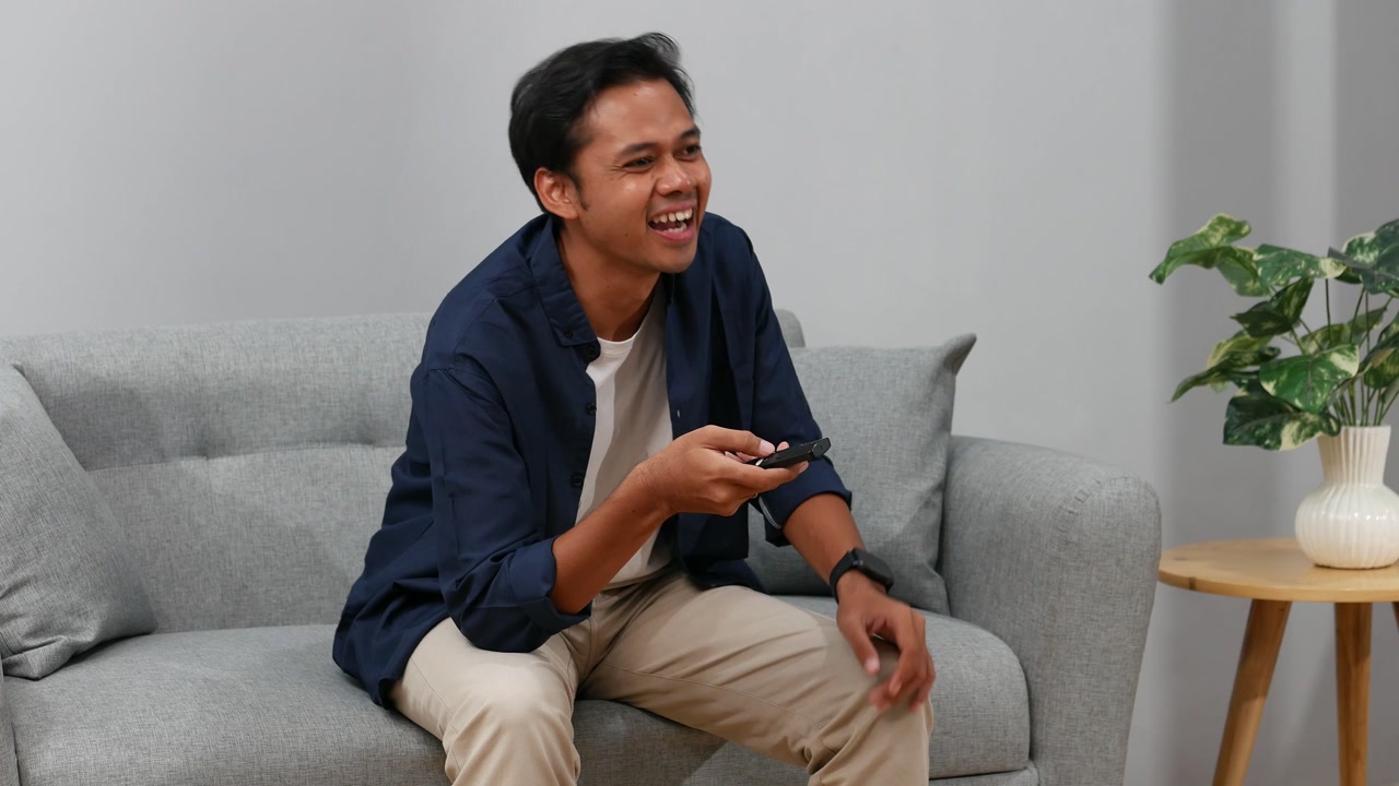 Glad ung asiatisk mand sidder på sofaen med fjernbetjening og ser tv derhjemme. Mennesker og fritidskoncept.