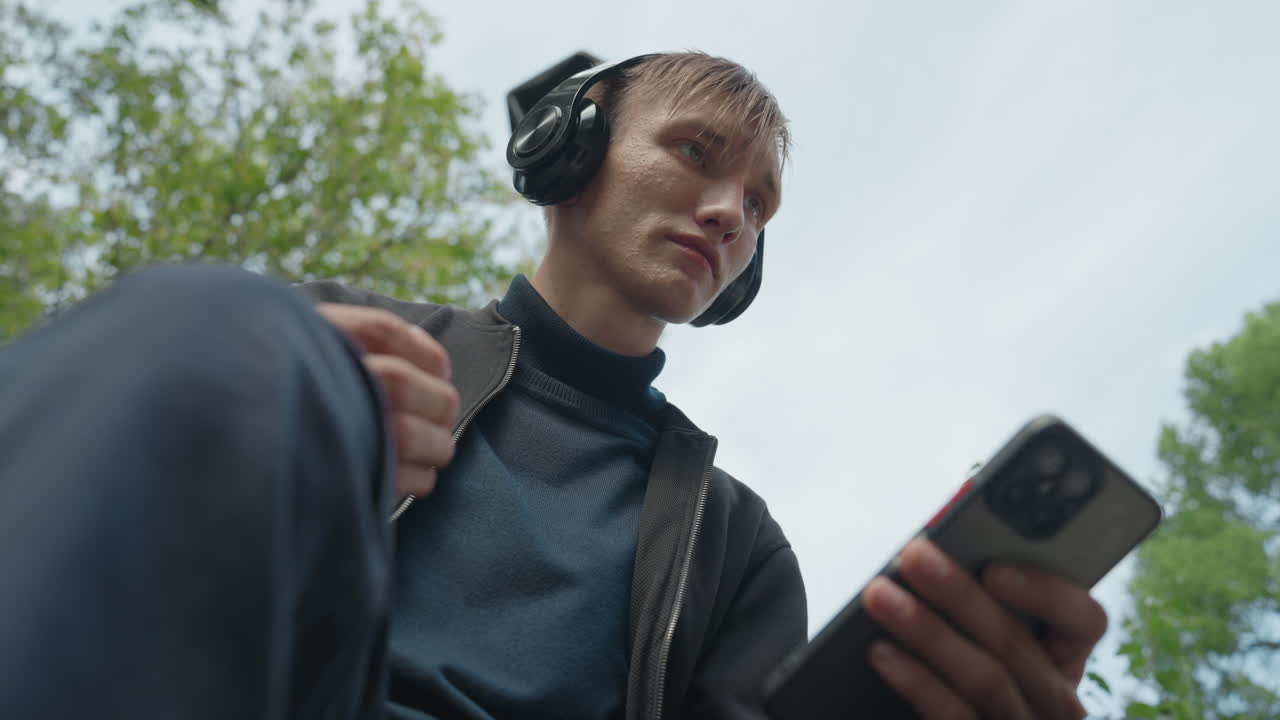 Uomo bianco che ascolta con le cuffie, telefono in basso nell'inquadratura, musicista di strada che cattura una registrazione sul campo e modifica le impostazioni, maglione e giacca blu, prospettiva dal basso con alberi e cielo sullo sfondo