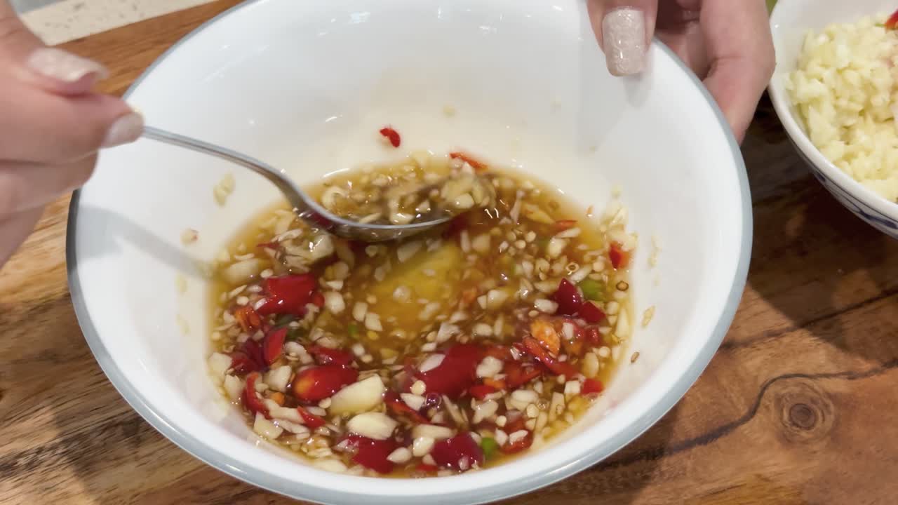 A hand stirs chili, garlic, and fish sauce in a white bowl on a wooden table under bright lighting, with smooth camera movement and close-up framing