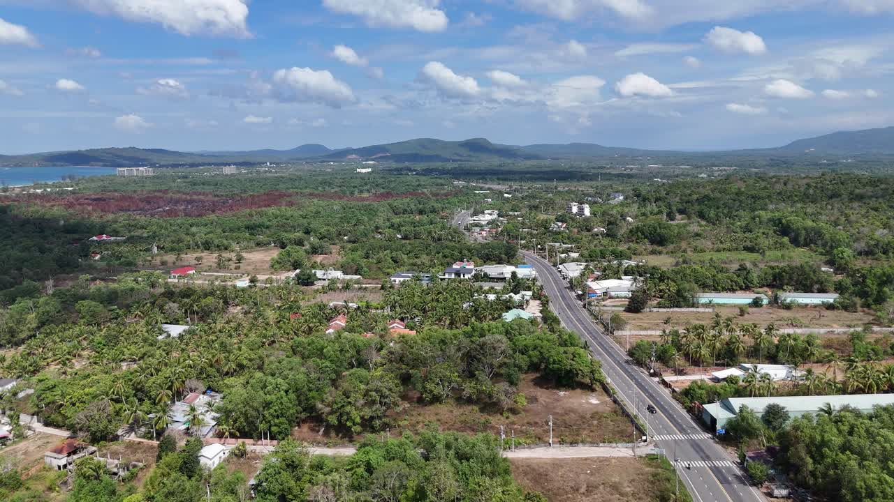 Aerial footage reveals a main road with less traffic, greenery bordering each side, and occasional glimpses of residents, creating a serene and inhabited streetscape. Passing clouds and their shadow