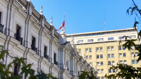 chilean flag swaying on top of the presidential palace, la moneda facade, tree branches in foreground, chile