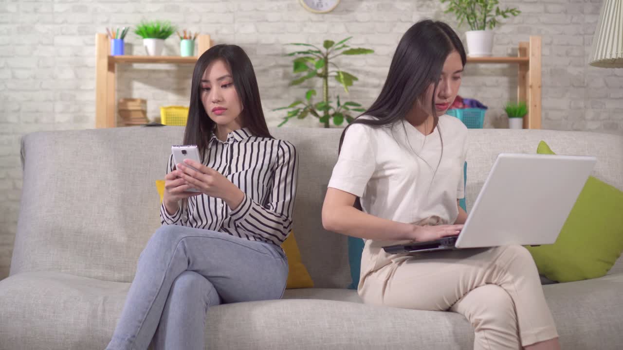 두 명의 젊은 아시아 여성이 거실의 소파에 앉아 기기를 사용하고 있습니다.