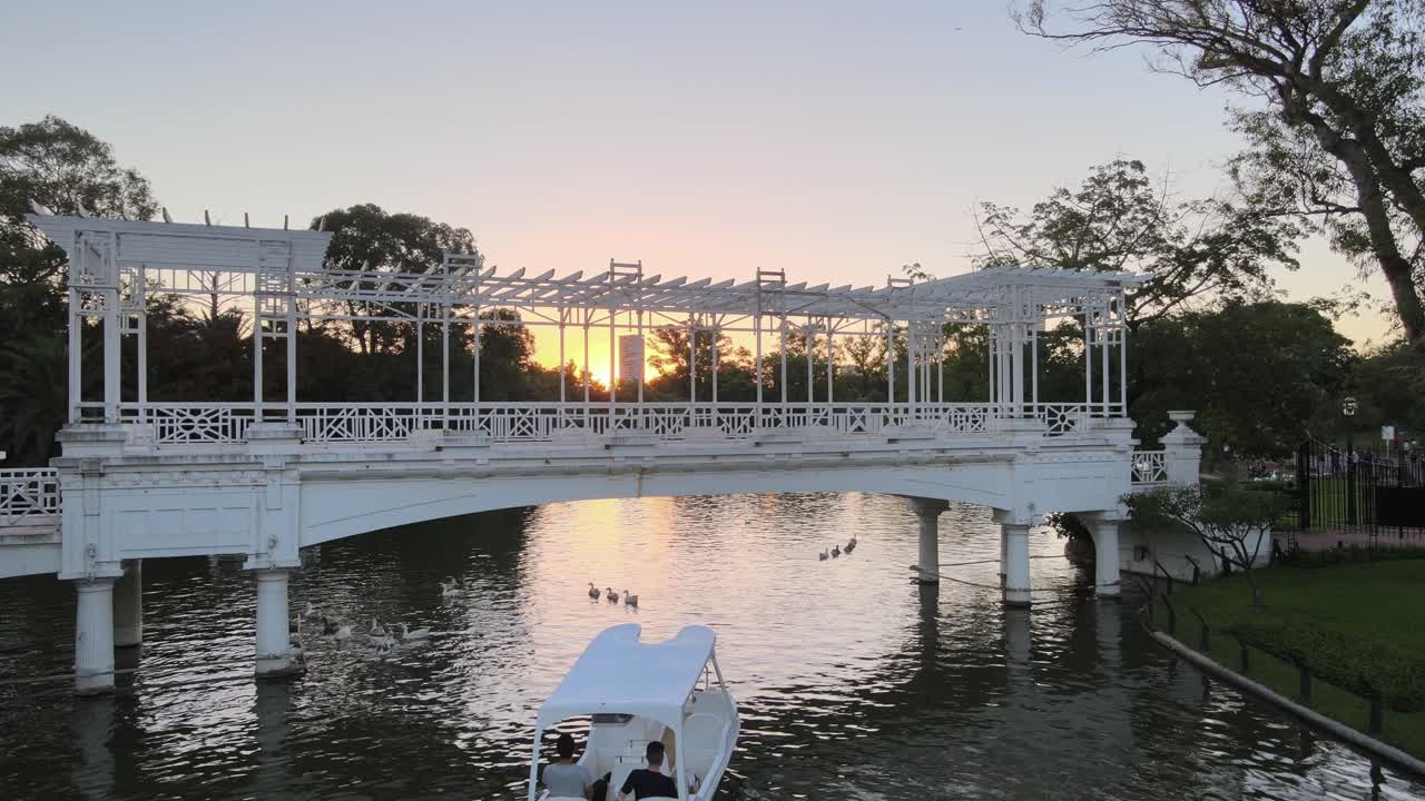 Aerial pan right of boat sailing under white bridge in Rosedal gardens near Palermo Woods at sunset, Buenos Aires