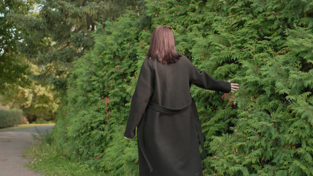 어린 여성 관광객이 어두운 겨울 코트를 입고 부드러운 햇빛 아래 고요한 공원 산책로를 따라 초록색 잎을 만지며, 손으로 울타리를 가로지르는 뒷모습이에요. 자연 여행 분위기가 느껴져요.