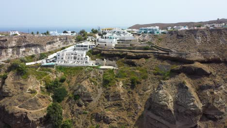 Aerial-descending-over-Greek-Island,-rocky-cliff-with-villas-and-fishing-boats-in-the-Mediterranean-Sea-on-beach-in-Santorini,-Greece