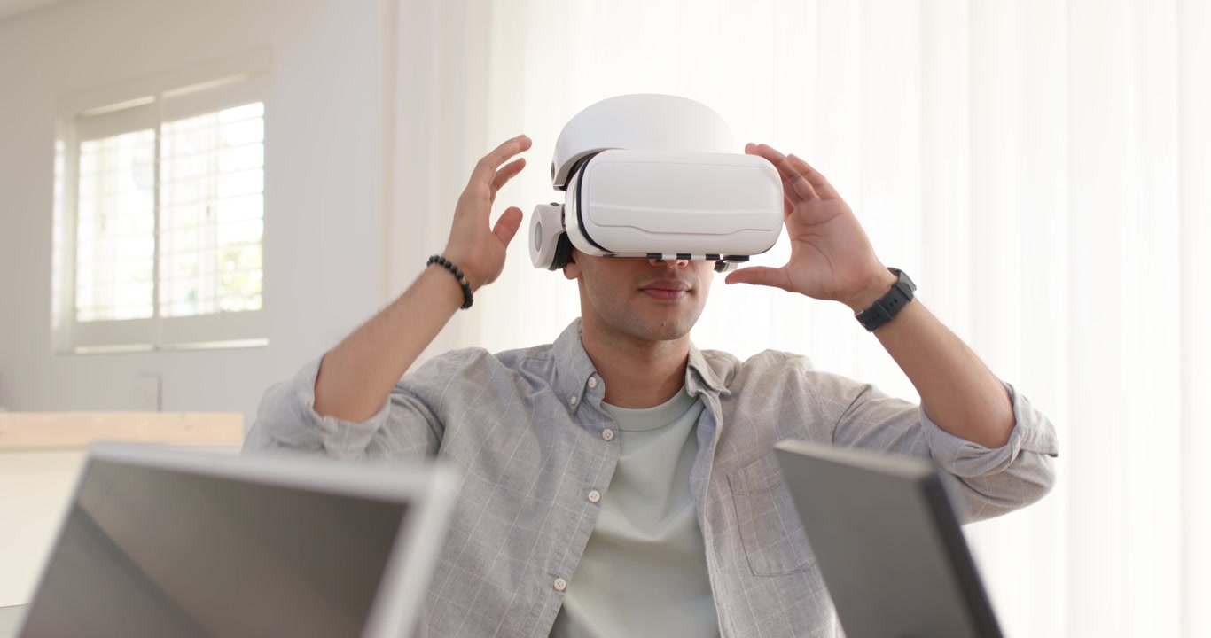 Uomo che solleva un visore VR bianco da una scrivania di vetro in un ufficio domestico, indossandolo sugli occhi per esplorare la scena