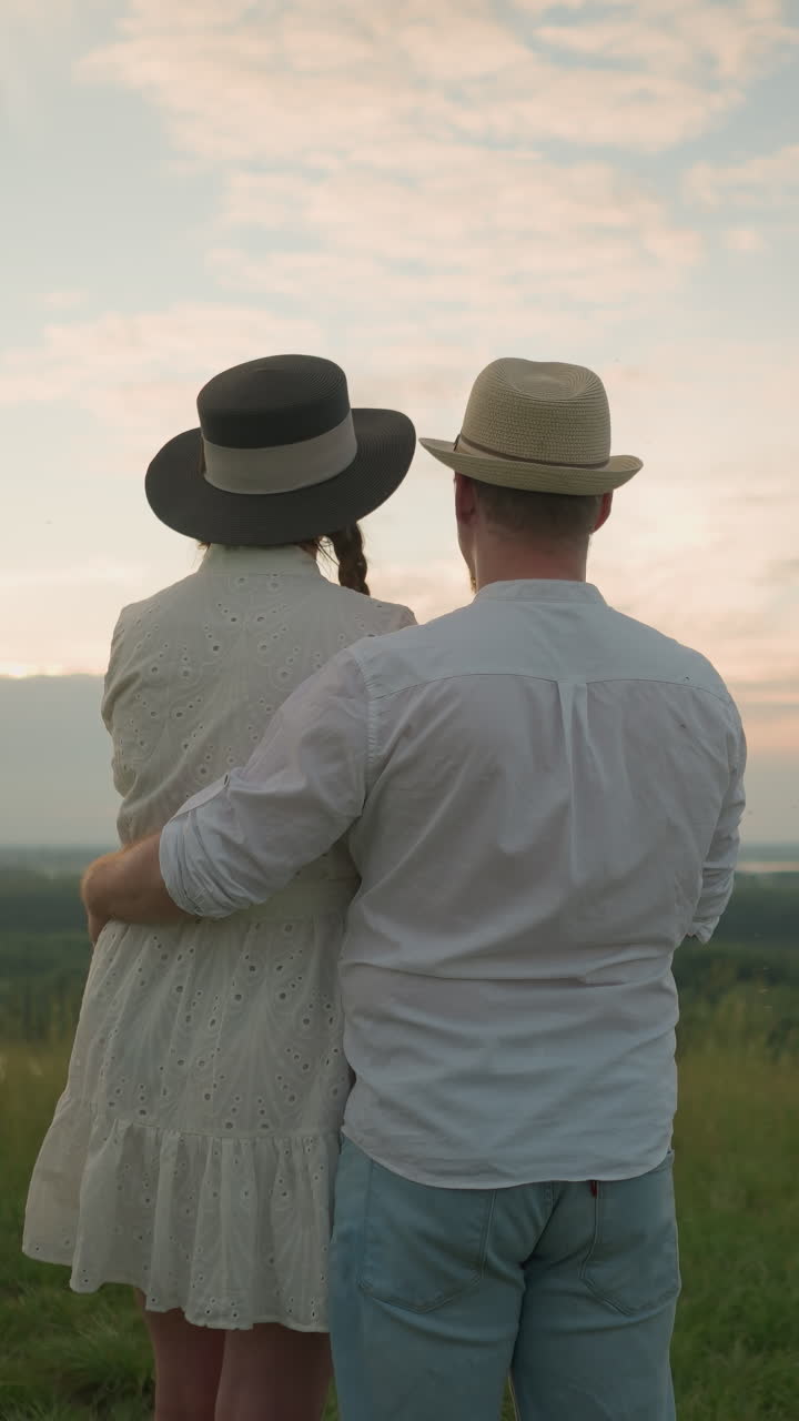 A close-up of a couple standing on a grassy hill at sunset, gazing at the sky. The man, in a white shirt, hat, and jeans, gently holds the woman by the waist. She wears a black hat and a white dress