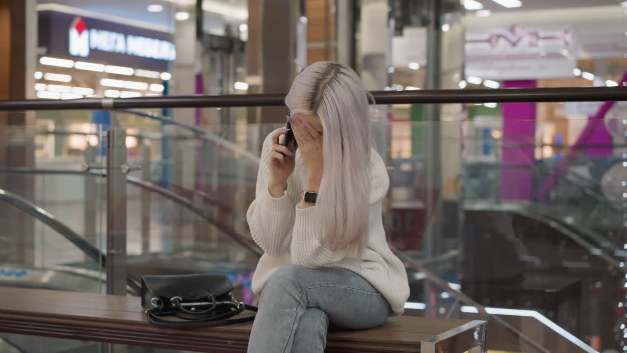stylish woman seated on mall bench with phone held to ear, eyes closed and posture slumped in exhaustion, dressed in sweater, denim jeans and boots, purse beside her, lights and glass railing