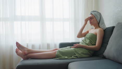 woman sitting on couch with towel around body and head gently rubs cucumber on forehead as part of relaxed self-care routine, serene environment enhanced by bright natural light