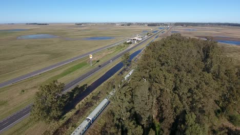 aerial view of a train cruising alongside a highway in a rural landscape, clear skies in the outskirts of buenos aires