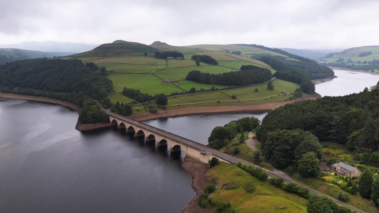 Drone footage glides over an arched stone bridge spanning a wide reservoir, surrounded by lush green hills and forests under overcast skies