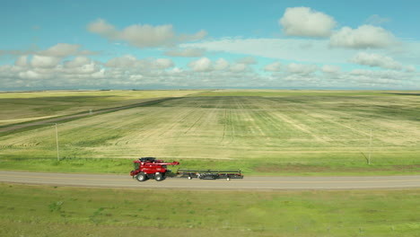 modern red combine harvester versatile machines drive on dirt roads by expansive flat green farmland of crops on sunny day, saskatchewan, canada, above aerial sideways