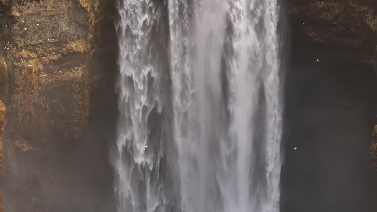 Aerial close-up of Skógafoss waterfall in Iceland, as glacial water plunges down dark cliffs with immense force, creating mist and dramatic textures on the rock walls.