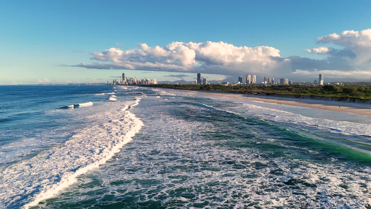 Drone footage captures Gold Coast's shoreline with waves, beachgoers, and city skyline under clear skies and vibrant lighting