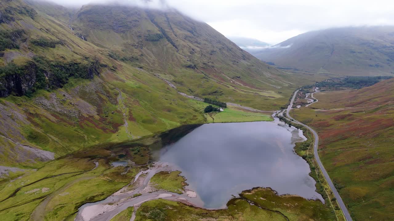Aerial drone footage of Loch Achtriochtan and road A82 in the Glencoe mountain range in the Scottish Highlands, taken in summer on a clear but misty day, morning time.