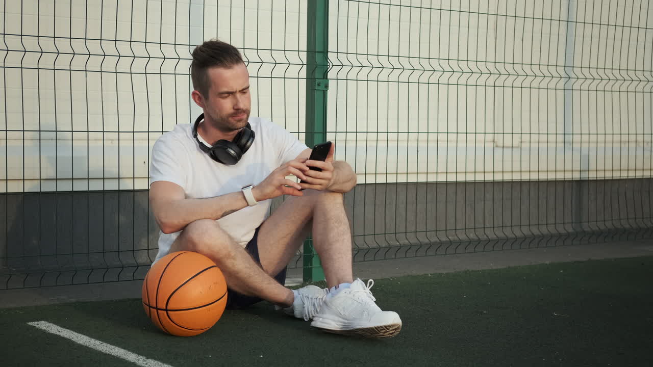 농구장 에 앉아 있는 남자 가 전화 를 보고 있다