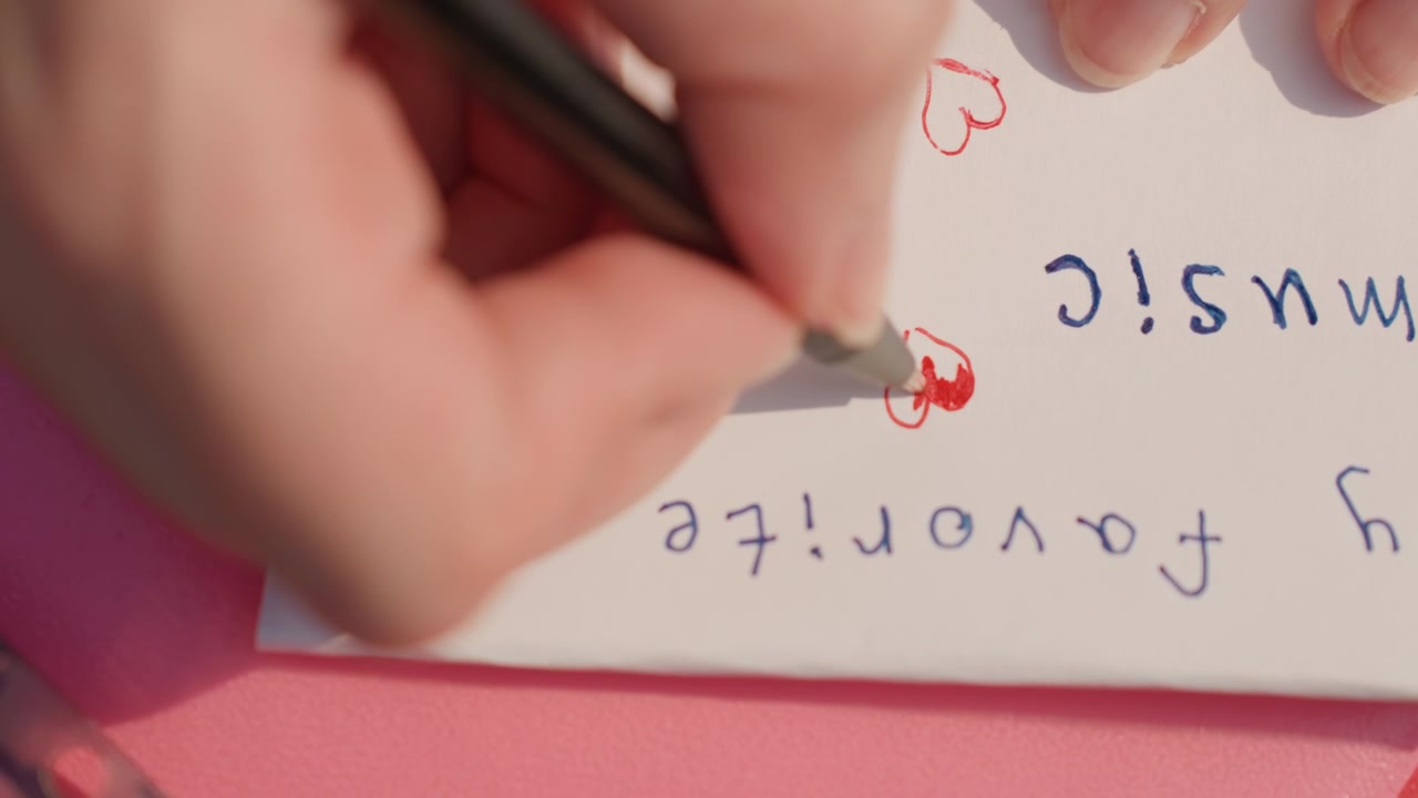 Hand holding pen draws red hearts on paper with handwritten text My favorite music in close up view. scene focuses on precise movement of drawing small details and adding elements
