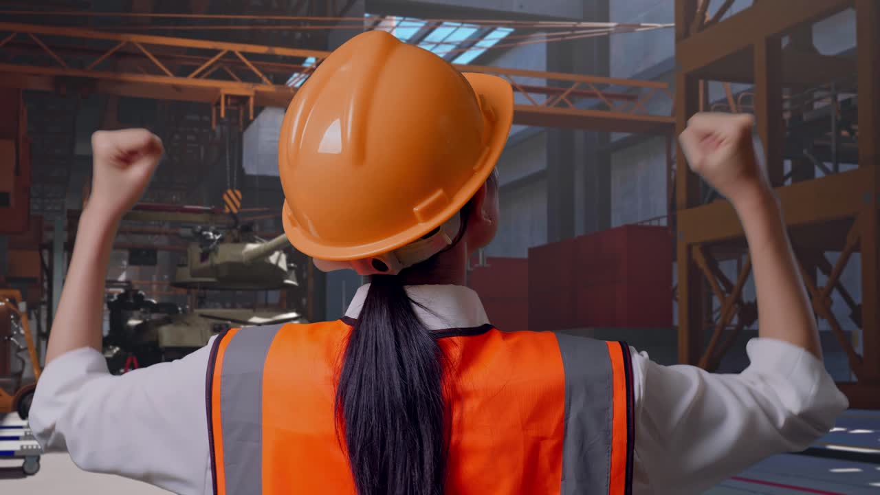 Primo piano da dietro di un'ingegnera con il casco di sicurezza che alza le mani per festeggiare mentre lavora, carro armato militare in costruzione in uno stabilimento industriale
