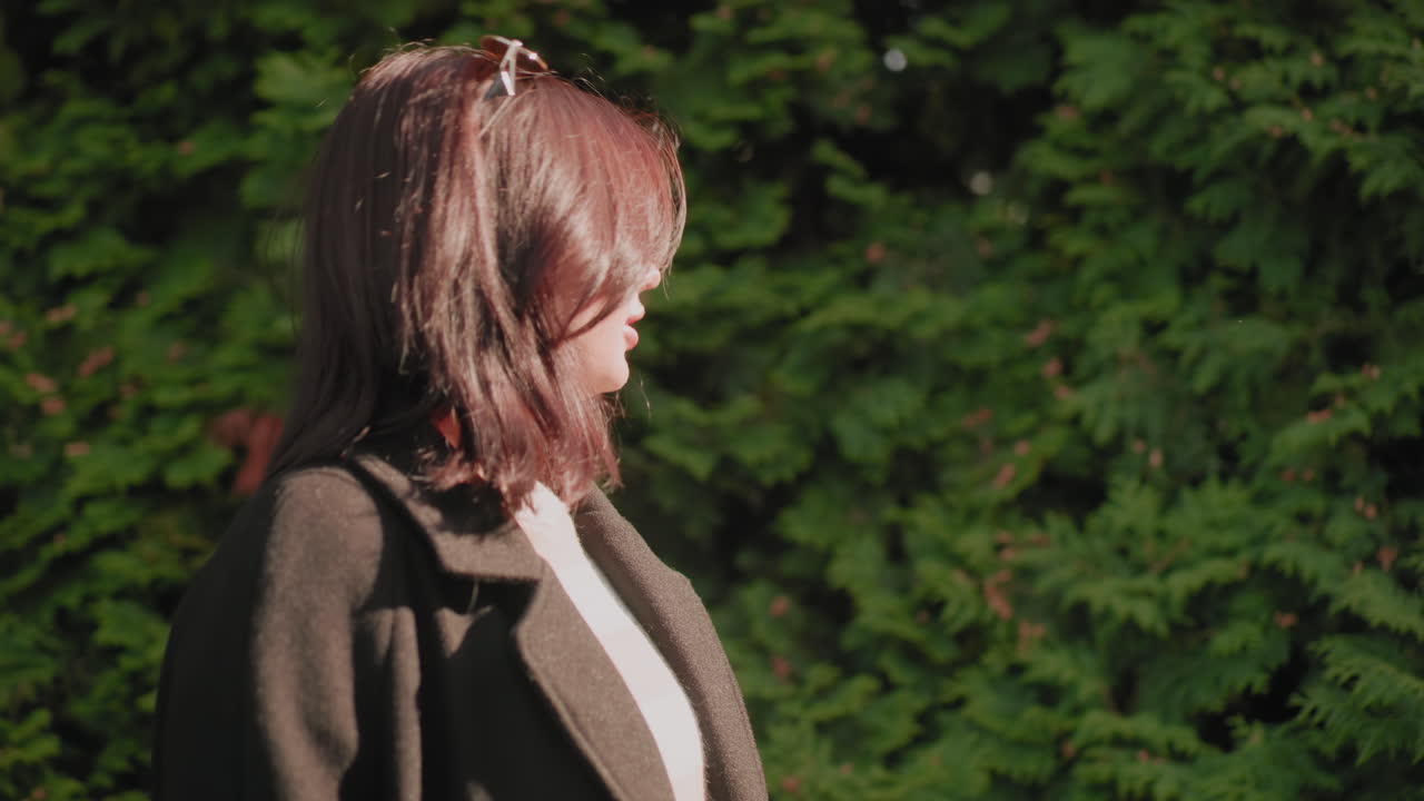 세련된 여성이 초록색 울타리 옆을 거닐면서 손으로 나뭇잎을 스치고, 긴 코트를 입고, 부드러운 햇살 아래 차분한 분위기로 자연 속 오솔길을 따라 여유롭게 산책하는 측면 모습이에요. 싱그러운 계절의 색깔이 돋보이는 평온한 야외 순간이죠.