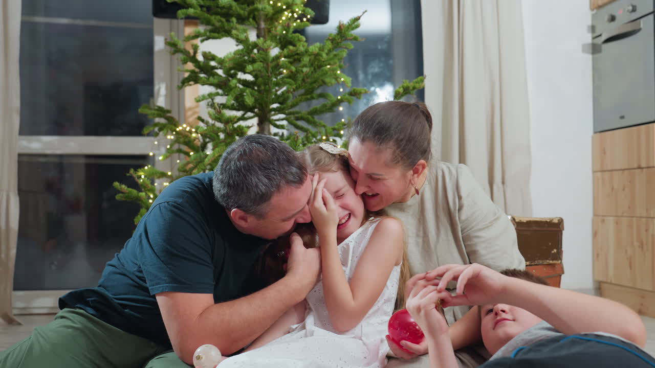 Father and mother lovingly kissing young daughter on both cheeks while smiling son lies relaxed in front of them holding colorful christmas ornament during festive holiday season near decorated tree