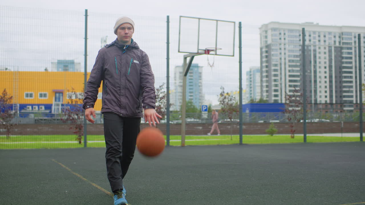 Uomo che palleggia con un pallone da basket su un campo all'aperto. Parco urbano nuvoloso con grattacieli sullo sfondo, canestro recintato sul bordo del campo, giocatore che indossa una giacca imbottita e un berretto di lana, scarpe da ginnastica che battono.