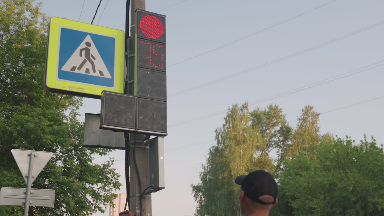 Pedestrian Man Waiting At Red Signal, Rear View With Cap And Backpack Straps, Looking Toward Pole Mounted Light, Trees And Wires Backdrop, Suburban Intersection, Tense Pause, Anxious Body Language